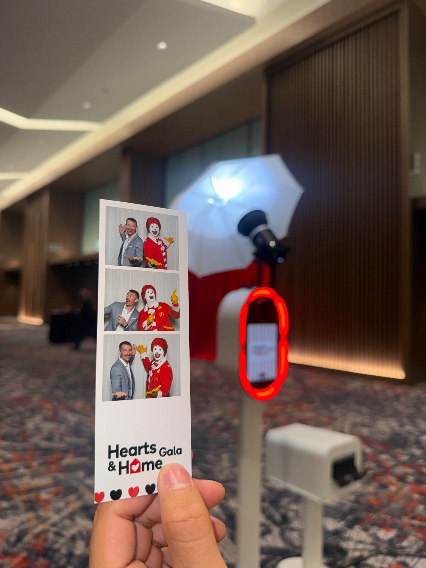 A hand holding up a Memento photo strip of three posed photos, with the photo booth glowing in the background of a gala ballroom